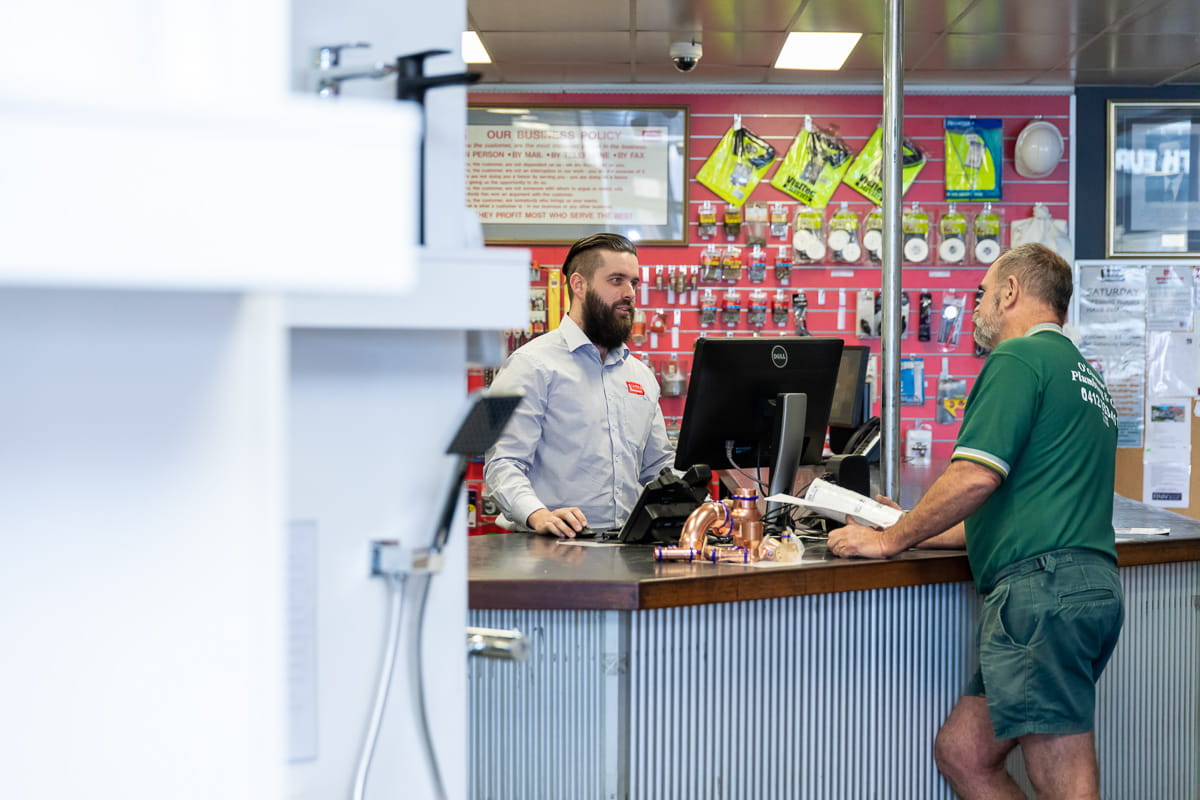 A long shot of a Galvins employee serving a tradesperson at the sales counter with products behind them on wall and showerheads on display in foreground