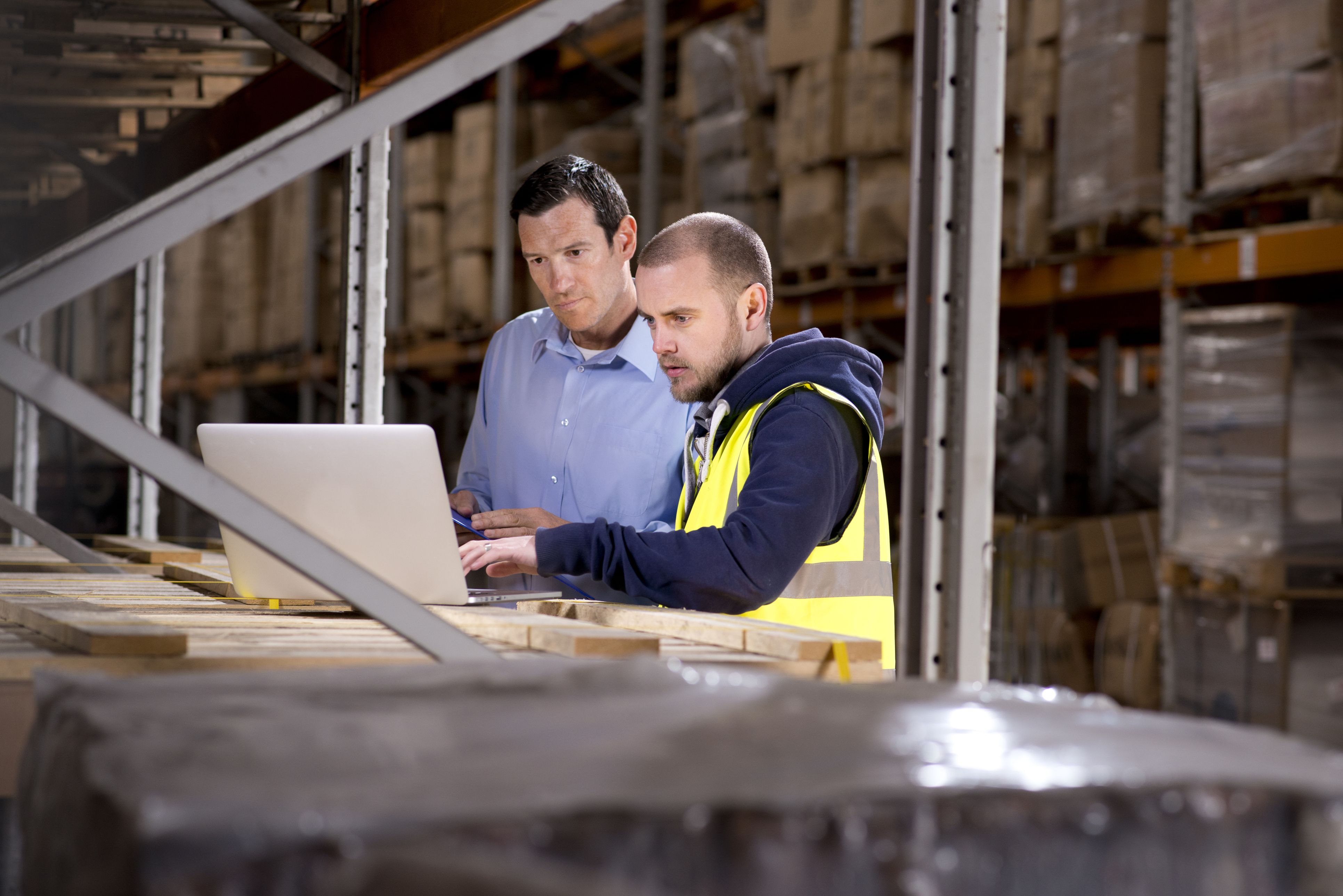 Two men in a warehouse looking at a laptop together - one is in PPE and the other in business attire